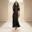 Woman in a black dress with gold jewelry standing in a bright room.