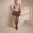 Woman in a brown pleated skirt and knee-high boots standing in a softly lit room.