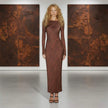 Woman in a long brown dress standing between two large textured paintings.