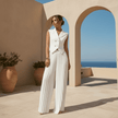 Woman in a white outfit standing in front of a beige wall with arches and potted plants, with a blue sky and ocean in the background.