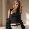 Woman wearing a black one-shoulder top and white pants in a softly lit room.