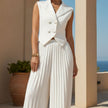 Person wearing a white sleeveless top and pleated skirt standing against a beige wall with a plant and ocean view.