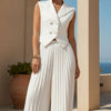 Person wearing a white sleeveless top and pleated skirt standing against a beige wall with a plant and ocean view.