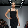 Model wearing a black NORVANE Riviera Bandage Dress on a fashion runway with an audience.