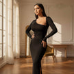 Woman in a black dress standing in a sunlit room with wooden floors and white walls.